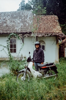 An individual wearing a helmet and jacket is standing next to a motorcycle in front of an old, weathered house with a tiled roof. The house appears aged and somewhat deteriorated, with plants growing around it. The ground is covered in tall grass, indicating a somewhat neglected area.