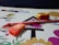 A drawing book lies open with a colorful cover featuring cartoon birds and trees. An orange eraser with branding is placed on the book, alongside a yellow and red sharpener and an orange pencil. The background is a dark surface.