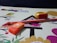 A drawing book lies open with a colorful cover featuring cartoon birds and trees. An orange eraser with branding is placed on the book, alongside a yellow and red sharpener and an orange pencil. The background is a dark surface.