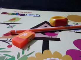 A drawing book lies open with a colorful cover featuring cartoon birds and trees. An orange eraser with branding is placed on the book, alongside a yellow and red sharpener and an orange pencil. The background is a dark surface.