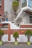 A residential building facade featuring a staircase with a metal railing, adorned with potted plants. The stairs lead to a landing area with additional potted plants and a blue plastic chair. The exterior walls are a combination of red brick and light gray material. Several green potted shrubs are lined up along the street in light green planters.