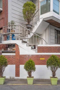A residential building facade featuring a staircase with a metal railing, adorned with potted plants. The stairs lead to a landing area with additional potted plants and a blue plastic chair. The exterior walls are a combination of red brick and light gray material. Several green potted shrubs are lined up along the street in light green planters.