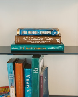 A collection of books is stacked neatly on two levels of a glass shelf. On the top shelf, three books are stacked horizontally. The titles include 'Starfarers' by Poul Anderson, 'All Cloudless Glory,' and 'U.S. Battleships.' The spines of additional books can be seen on the bottom level, including titles like 'AMBRA' and '1993 Directory of Members.' The books have varied colored covers, mostly in blue, green, and brown tones.
