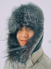 A person is wearing a warm fur hat and a jacket suitable for cold weather. Their face is partly obscured by the hood's fur trim. Snowflakes appear to be gently falling around them, and their skin tone is slightly flushed from the cold.