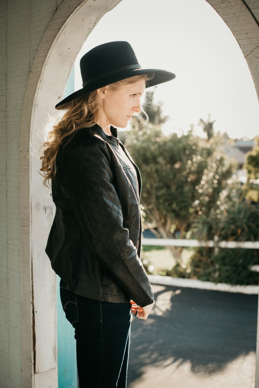 woman in black leather jacket and black hat standing beside white wall during daytime