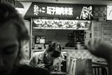 A person is seated at a fast-food restaurant, engaging in a meal. The backdrop features a brightly lit menu board displaying dishes and written in an East Asian script. The black and white filter adds a nostalgic or moody ambiance, with another person's silhouette visible in the foreground.