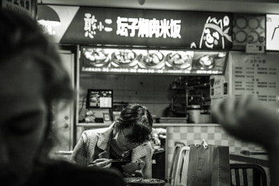 A person is seated at a fast-food restaurant, engaging in a meal. The backdrop features a brightly lit menu board displaying dishes and written in an East Asian script. The black and white filter adds a nostalgic or moody ambiance, with another person's silhouette visible in the foreground.