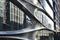 A modern architectural facade with smooth, curved metallic surfaces. Large reflective glass panels mirror the surrounding brick buildings, creating a dynamic interplay between different architectural styles.