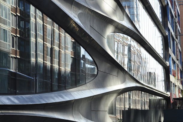 A modern architectural facade with smooth, curved metallic surfaces. Large reflective glass panels mirror the surrounding brick buildings, creating a dynamic interplay between different architectural styles.