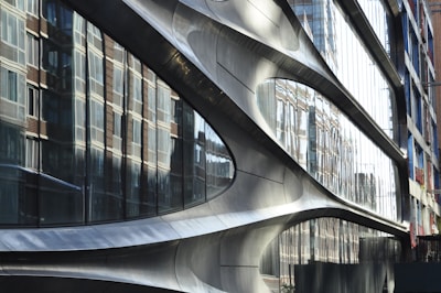 A modern architectural facade with smooth, curved metallic surfaces. Large reflective glass panels mirror the surrounding brick buildings, creating a dynamic interplay between different architectural styles.