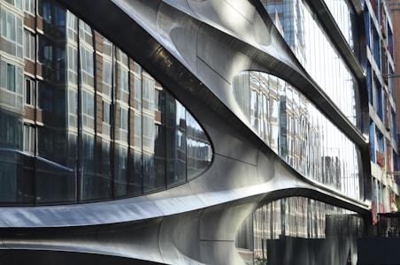 A modern architectural facade with smooth, curved metallic surfaces. Large reflective glass panels mirror the surrounding brick buildings, creating a dynamic interplay between different architectural styles.