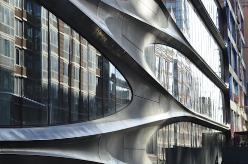 A modern architectural facade with smooth, curved metallic surfaces. Large reflective glass panels mirror the surrounding brick buildings, creating a dynamic interplay between different architectural styles.