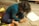 A young child with curly hair sits on a wooden floor, intensely focused on drawing with colored pencils and crayons. The surrounding area has a colorful rug and scattered art supplies, including paper and a box of crayons.