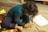 A young child with curly hair sits on a wooden floor, intensely focused on drawing with colored pencils and crayons. The surrounding area has a colorful rug and scattered art supplies, including paper and a box of crayons.
