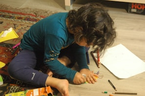 A young child with curly hair sits on a wooden floor, intensely focused on drawing with colored pencils and crayons. The surrounding area has a colorful rug and scattered art supplies, including paper and a box of crayons.