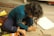 A young child with curly hair sits on a wooden floor, intensely focused on drawing with colored pencils and crayons. The surrounding area has a colorful rug and scattered art supplies, including paper and a box of crayons.