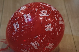 A red balloon featuring white text and patterns, including the phrase 'Happy Birthday' and festive confetti and streamers, set against a wooden floor.