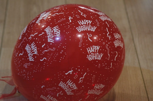 A red balloon featuring white text and patterns, including the phrase 'Happy Birthday' and festive confetti and streamers, set against a wooden floor.