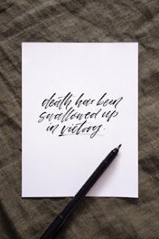 A sheet of white paper with the phrase 'death has been swallowed up in victory' written in elegant black calligraphy. The paper rests on a textured dark green fabric background. A black pen is positioned diagonally on the paper.