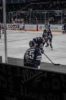 A hockey game is taking place on the ice rink with players in action. Several athletes in blue uniforms are participating, while one player in white and red gear is visible in the background sitting near the goal post. The arena has a dark seating area with a few spectators.