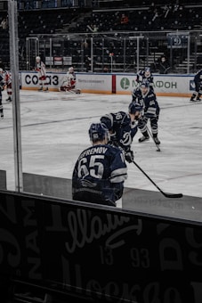 A hockey game is taking place on the ice rink with players in action. Several athletes in blue uniforms are participating, while one player in white and red gear is visible in the background sitting near the goal post. The arena has a dark seating area with a few spectators.