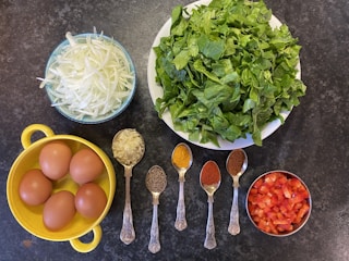 A variety of fresh and prepared ingredients laid out on a dark surface. The arrangement includes a large bowl of leafy green spinach, thinly sliced white onions in a blue bowl, and chopped red bell peppers in a small silver bowl. A yellow container holds four brown eggs. Five ornate silver spoons are filled with different spices: minced garlic, cumin seeds, turmeric, red chili powder, and ground coriander.