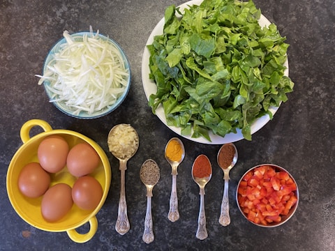 A variety of fresh and prepared ingredients laid out on a dark surface. The arrangement includes a large bowl of leafy green spinach, thinly sliced white onions in a blue bowl, and chopped red bell peppers in a small silver bowl. A yellow container holds four brown eggs. Five ornate silver spoons are filled with different spices: minced garlic, cumin seeds, turmeric, red chili powder, and ground coriander.