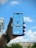 A hand is holding a smartphone in front of a blue sky with clouds. The phone screen shows an airplane flying directly overhead, creating an illusion that the plane is part of the sky view.