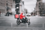 A food delivery rider wearing a bright red jacket and helmet rides a black scooter through a city street. The background shows motion blur indicating speed, with blurred outlines of buildings and other vehicles.