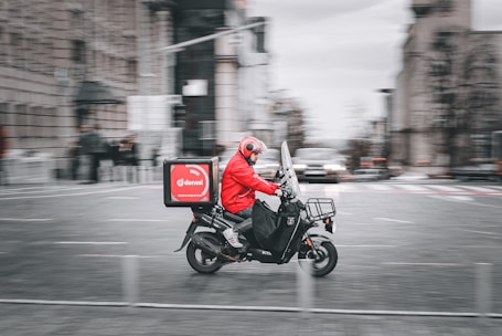 A food delivery rider wearing a bright red jacket and helmet rides a black scooter through a city street. The background shows motion blur indicating speed, with blurred outlines of buildings and other vehicles.