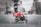 A food delivery rider wearing a bright red jacket and helmet rides a black scooter through a city street. The background shows motion blur indicating speed, with blurred outlines of buildings and other vehicles.