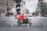 A food delivery rider wearing a bright red jacket and helmet rides a black scooter through a city street. The background shows motion blur indicating speed, with blurred outlines of buildings and other vehicles.