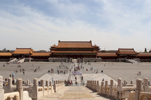 A vast, open courtyard leads to a series of traditional Chinese palatial structures with ornate red and gold roofs. The scene is bustling with tourists, who meander along the wide stone pathways. The sky is a clear blue, providing a serene backdrop to the historic architecture.
