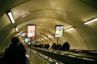 A long indoor escalator surrounded by a metallic tunnel, with people, mostly in dark clothing, riding both up and down. Advertising boards are positioned along the pathway, displaying colorful posters. The tunnel is illuminated by linear fluorescent lights attached to the curved ceiling.