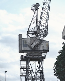 A large, industrial crane with a metal lattice structure towering against a cloudy sky. The crane features a control cabin with a sign that reads 'Stothert & Pitt Ltd, Bath, England'. The overall structure has a weathered and aged appearance, suggesting it is part of an old or unused dockyard or industrial site.