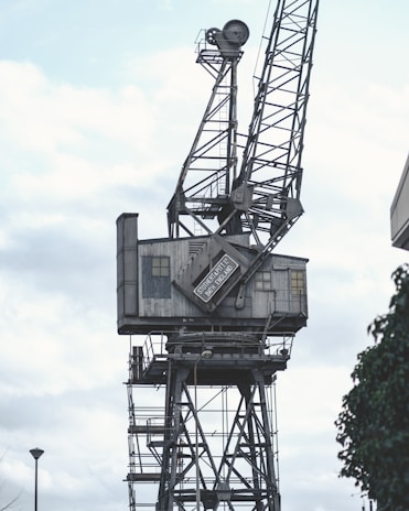A large, industrial crane with a metal lattice structure towering against a cloudy sky. The crane features a control cabin with a sign that reads 'Stothert & Pitt Ltd, Bath, England'. The overall structure has a weathered and aged appearance, suggesting it is part of an old or unused dockyard or industrial site.