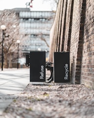 Two black waste bins labeled 'Refuse' and 'Recycle' are positioned side by side on a sidewalk. They are placed against a high brick wall in an urban setting. The path is lined with bare trees and lampposts, and a modern building is visible in the background.