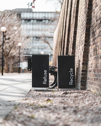 Two black waste bins labeled 'Refuse' and 'Recycle' are positioned side by side on a sidewalk. They are placed against a high brick wall in an urban setting. The path is lined with bare trees and lampposts, and a modern building is visible in the background.