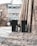 Two black waste bins labeled 'Refuse' and 'Recycle' are positioned side by side on a sidewalk. They are placed against a high brick wall in an urban setting. The path is lined with bare trees and lampposts, and a modern building is visible in the background.