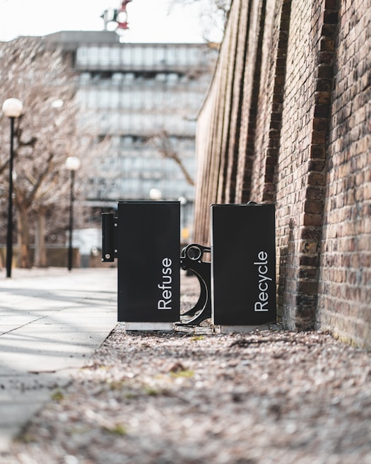 Two black waste bins labeled 'Refuse' and 'Recycle' are positioned side by side on a sidewalk. They are placed against a high brick wall in an urban setting. The path is lined with bare trees and lampposts, and a modern building is visible in the background.