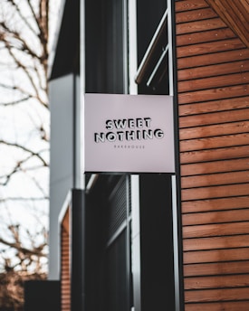 A bakery sign reading 'SWEET NOTHING Bakehouse' is mounted on the exterior of a modern building. The building features wooden siding and large windows. There are bare trees in the background.