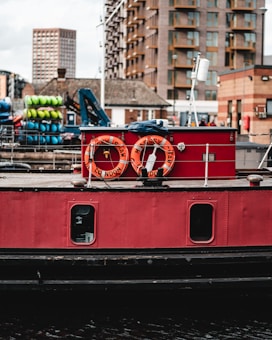 A red boat is moored in a harbor area, with life rings labeled 'Massey Shaw' and 'London' hanging on the side. Behind the boat, there are stacks of colorful kayaks or canoes, a crane, and modern buildings in the background, including a high-rise block.