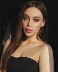 A young woman with long brown hair and green eyes is gazing directly at the camera. She is wearing a black strapless top and has subtle makeup with highlighted cheekbones and defined eyebrows.