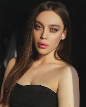 A young woman with long brown hair and green eyes is gazing directly at the camera. She is wearing a black strapless top and has subtle makeup with highlighted cheekbones and defined eyebrows.