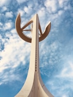 A tall white telecommunications tower with a futuristic design extends into a sky filled with scattered clouds. The structure features circular and pointed elements, contributing to its modern and sleek appearance.
