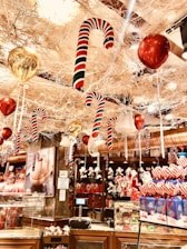 A festive store display is filled with holiday decorations, including large candy canes suspended from the ceiling, shiny red and gold balloons, and an assortment of Christmas-themed merchandise. The rich decor exudes a lively and vibrant atmosphere with colorful gift boxes and ornamented shelves.