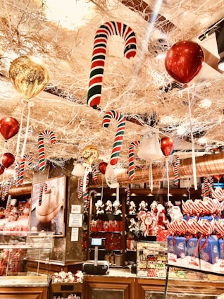 A festive store display is filled with holiday decorations, including large candy canes suspended from the ceiling, shiny red and gold balloons, and an assortment of Christmas-themed merchandise. The rich decor exudes a lively and vibrant atmosphere with colorful gift boxes and ornamented shelves.
