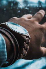 A close-up of a wrist adorned with a wooden watch, with a focus on the intricate details of the wristwatch and a blurred background through a glass window with water droplets.