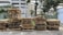 A collection of wooden pallets is neatly stacked on the side of an urban street. The pallets are arranged in columns, some taller than others, against a backdrop of a multi-story parking structure, trees, and a chain-link fence. There are green plants on the left and a tree overhanging on the right side of the scene.