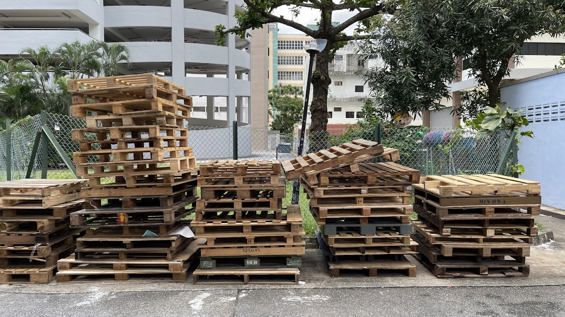 A collection of wooden pallets is neatly stacked on the side of an urban street. The pallets are arranged in columns, some taller than others, against a backdrop of a multi-story parking structure, trees, and a chain-link fence. There are green plants on the left and a tree overhanging on the right side of the scene.